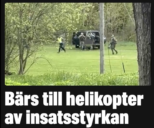 Grupp människor i uniform bär objekt mot svart fordon på gräsbevuxet fält, omgivet av träd. Text: "Bärs till helikopter av insatsstyrkan".