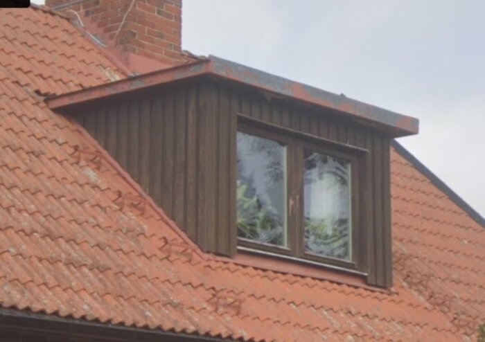 En takkupa med brunt trä och två fönster på ett tegeltak, i behov av renovering med ny panel och takplåt.