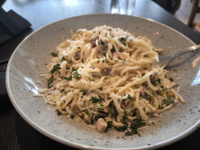Pasta Carbonara på en tallrik på Arlanda, toppad med parmesanost och örter, med en gaffel på sidan.