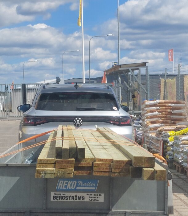 Bil med släpvagn fylld med brädor, parkerad vid Byggmax, klar för transport.