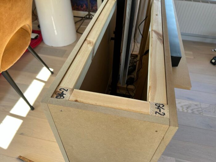 Låda av 45x45-reglar och 22 mm MDF med en inbyggd TV-hiss, öppen topp, planerad för trälock, inomhusmiljö, delar av möbler i bakgrunden.