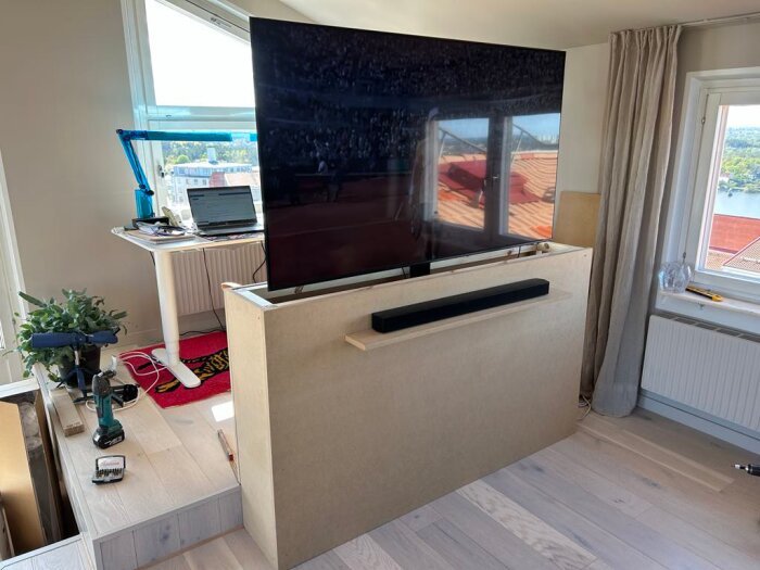 TV på hiss i en trälåda av MDF och 45x45-reglar, bredvid skrivbord med dator och verktyg i ett ljust rum med trägolv.