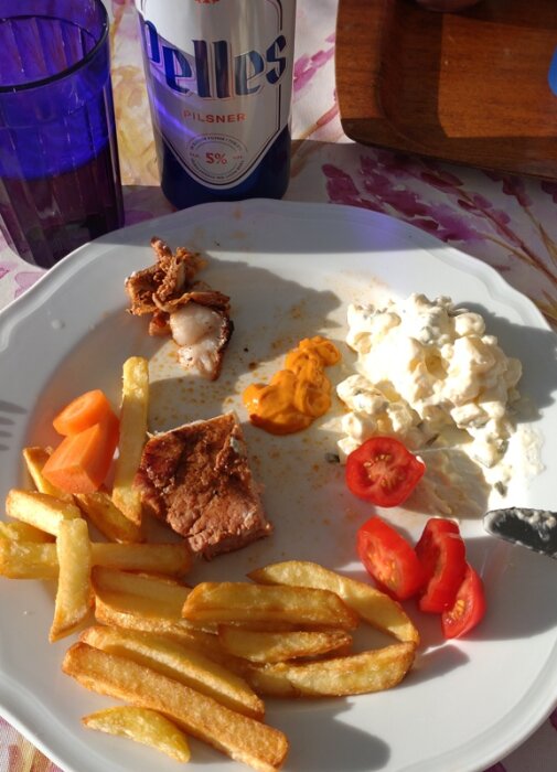 Tallrik med flintastek, pommes frites, potatissallad, tomater, morötter och senap. En flaska pilsner och ett glas på bordet.