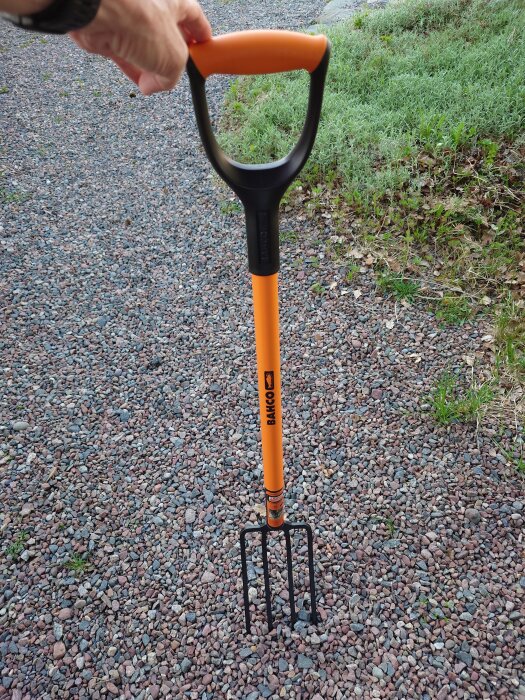 En hands hållande en grep med orange handtag på en grusgång, med grönt gräs vid sidan.