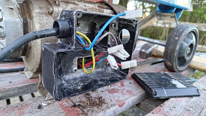 Trasig ellåda i glasfiber med färgade kablar fixade på en bänk, omgiven av verktyg och hjul.