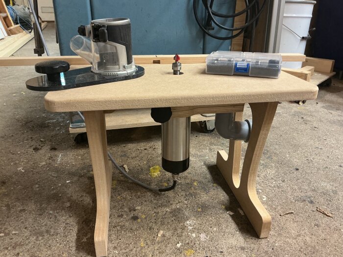 Fräsbord med frässpindel monterad, omgiven av verktyg och tillbehör i ett garage, byggd för hemmafix och renoveringsprojekt.