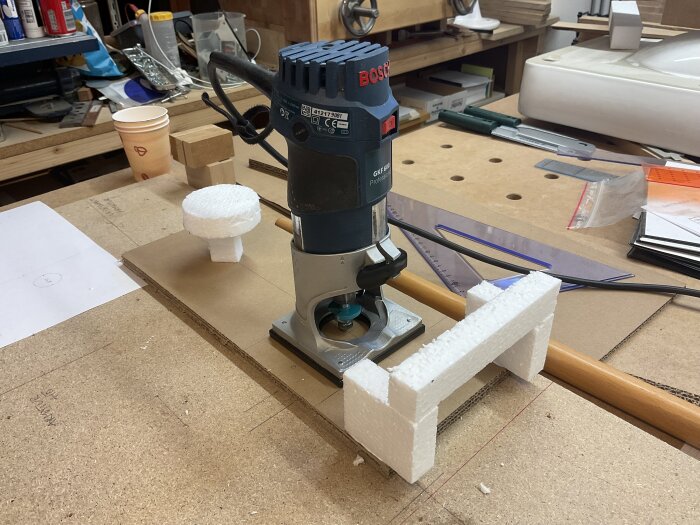 En fräsmaskin från Bosch på en arbetsbänk med olika verktyg och material runtomkring, inklusive skumplastdelar och papperskoppar.