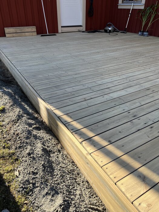 Nyrenoverad altan i trä efter slipning, visar en jämn yta intill en röd trävägg och en vit dörr. Verktyg syns vid kanten av bilden.