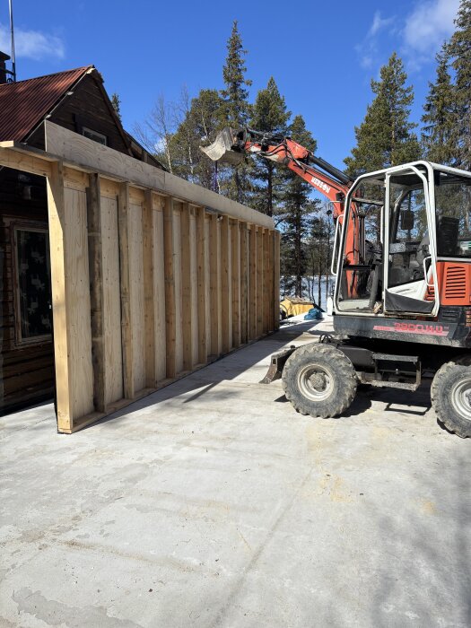 Träregelvägg under byggnad med grävmaskin i solsken, omgiven av träd.