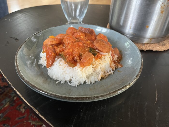 Het korvgryta på kokt ris serverad på en tallrik, med ett glas och gryta i bakgrunden.