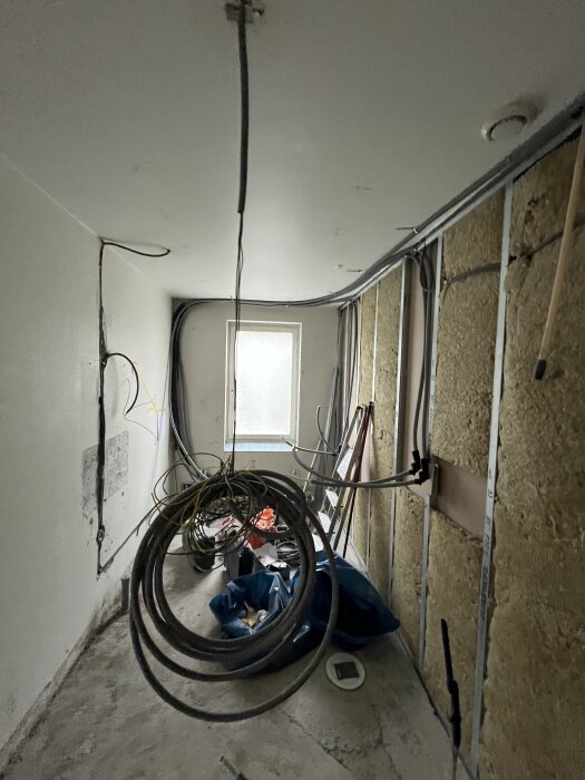 Badrum under renovering med blottade väggar, kablar hängande från taket och diverse verktyg och material utspridda på golvet.