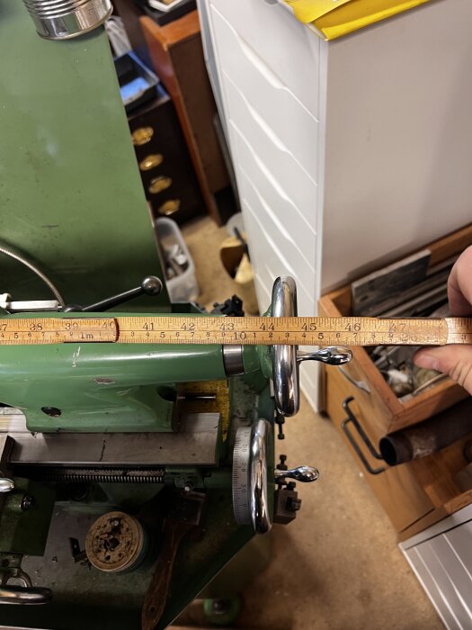 Gröngrön maskin med måttband ovanpå, omgiven av verktyg och möbler i verkstadsmiljö.