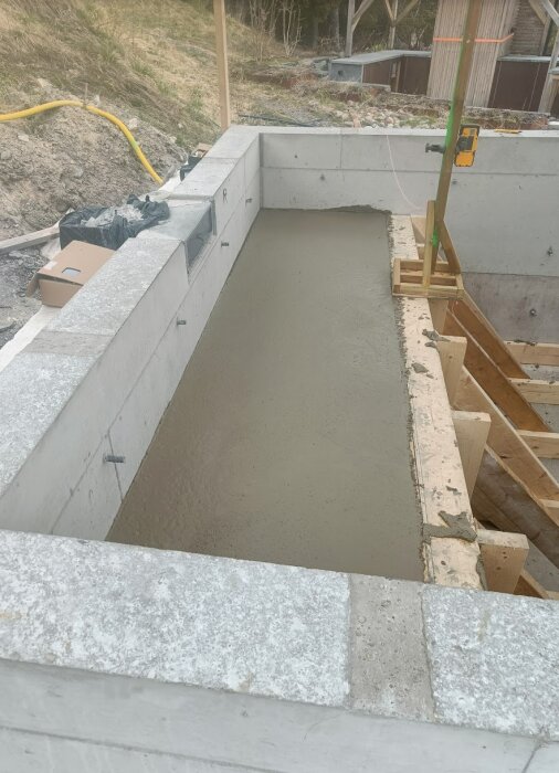 Betongpool under konstruktion med nyligen gjuten bänk längs ena sidan, omgiven av byggställningar och verktyg.