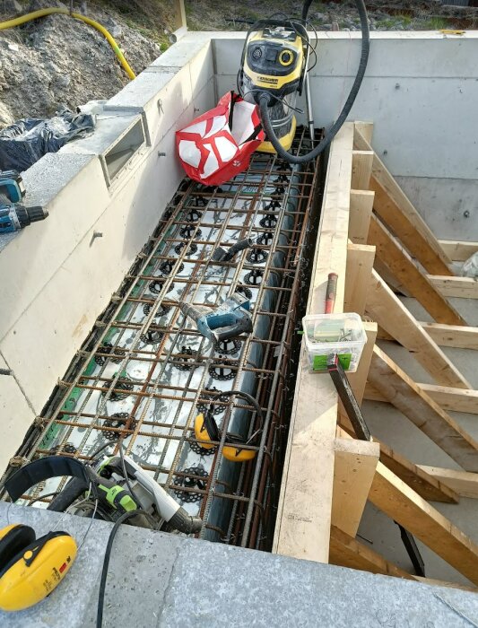 Byggarbetsplats med en gjuten pool under uppbyggnad, visar armeringsjärn, verktyg, en dammsugare och annan utrustning runt poolbänken.
