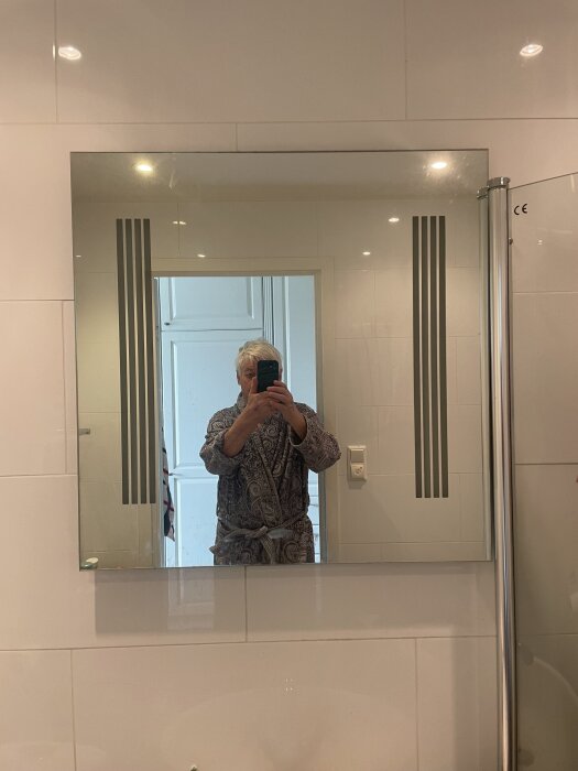 Badrumsspegel utan skåp med dekorativa ränder, person i morgonrock speglas när foto tas, fråga om byte av lysrör bakom spegeln.
