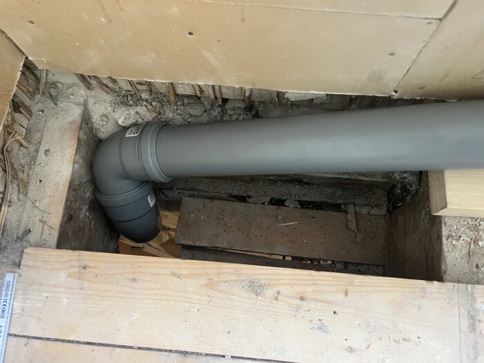 Grått avloppsrör med 87° och 45° böjar i träbjälklag, preparerat för installation bakom diskbänkskåp.