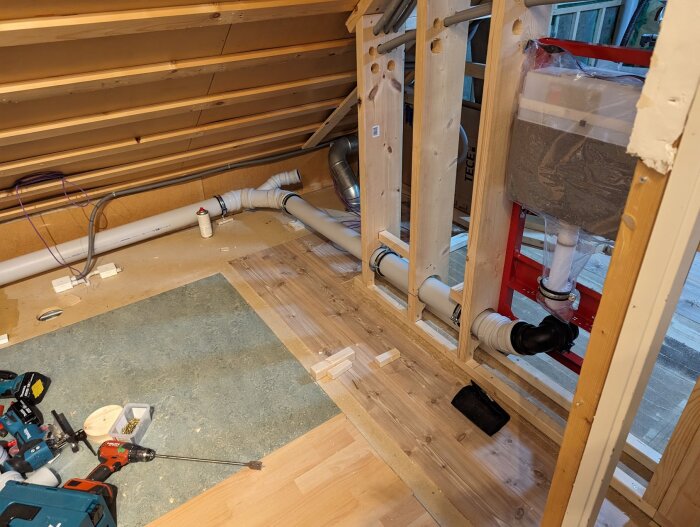 WC-fixtur och rörinstallation i ett bygge under arbete, visar träreglar, rör och verktyg i ett halvt färdigt badrum med exponerat material.