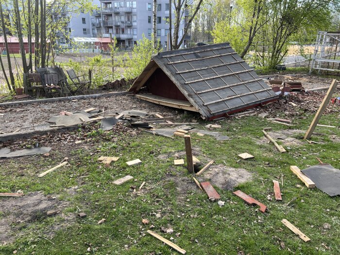 Ett halvt rivet tak ligger på marken, omgiven av byggmaterial och bråte i en trädgårdsmiljö med bostadshus i bakgrunden.