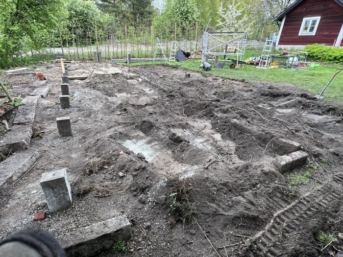 Grävd yta i trädgård med markförberedelse för byggprojekt, omgiven av växtlighet och byggmaterial.