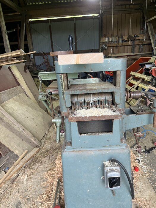 Gammal hyvelmaskin i ett träverkstad med träspån, brädbitar staplade runt och verktyg hängande på väggen.