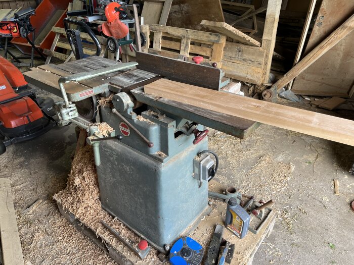 Gammal kombinationsmaskin i en verkstad, omgiven av spån och verktyg. En planka ligger på maskinen redo för hyvling.