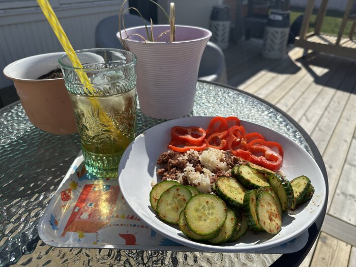 Stekt ris med hackad beyondburgare, skivad gurka och röd spetspaprika på en tallrik. Iste med sugrör i ett glas bredvid, på ett utebord.