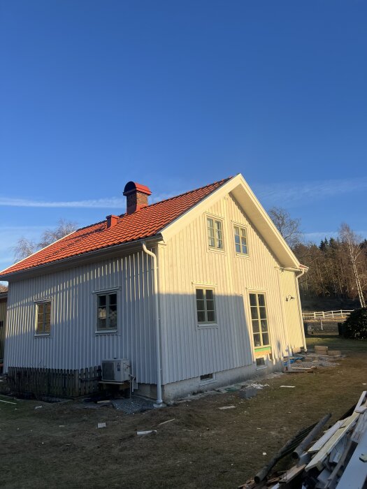 Fasadbyte på hus med vit panel och rött tak under klarblå himmel. Byggmaterial syns på marken.