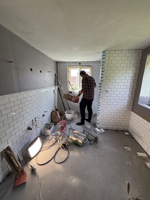 Pågående badrumsrenovering med en person, verktyg och material, vitt kakel på väggarna och ett fönster i bakgrunden.