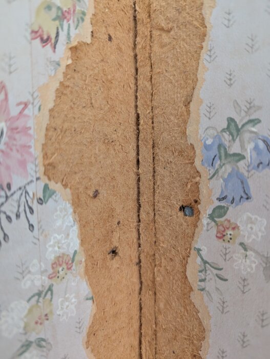 Riven tapet på vägg i gammal hälsingegård, synligt lager av brunt pappmaterial under blommönstrad yta.