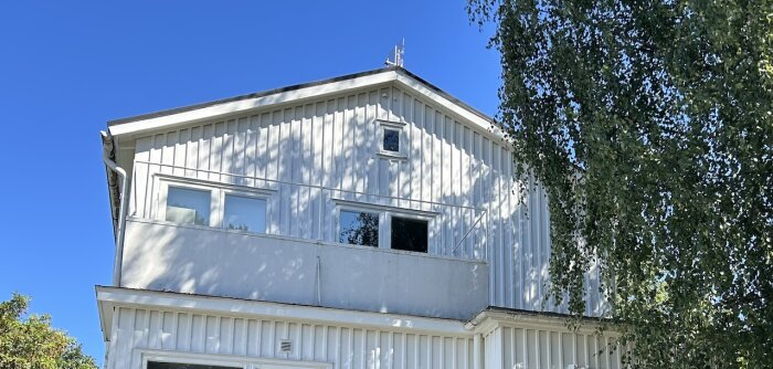Vit tvåvåningsvilla från 30-talet med platt takterrass. Solig dag med klarblå himmel och grönt träd till höger.