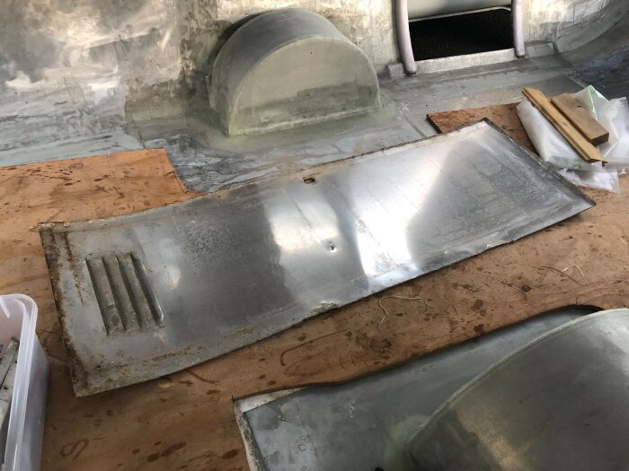 En gammal, bucklig aluminiumplåt med ventilationsgaller ligger på en arbetsbänk, omgiven av träspill och verktyg.