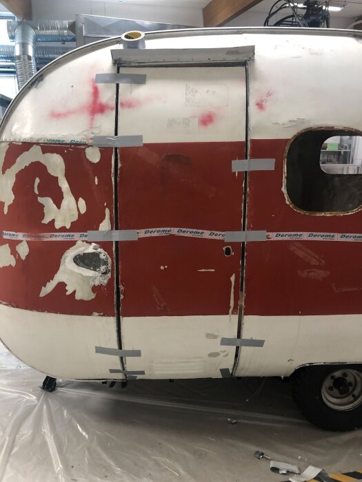 Husvagn med delvis röd och vit kaross där dörrplåten justeras, fäst med silvertejp och stöd för att passa in korrekt under renovering.