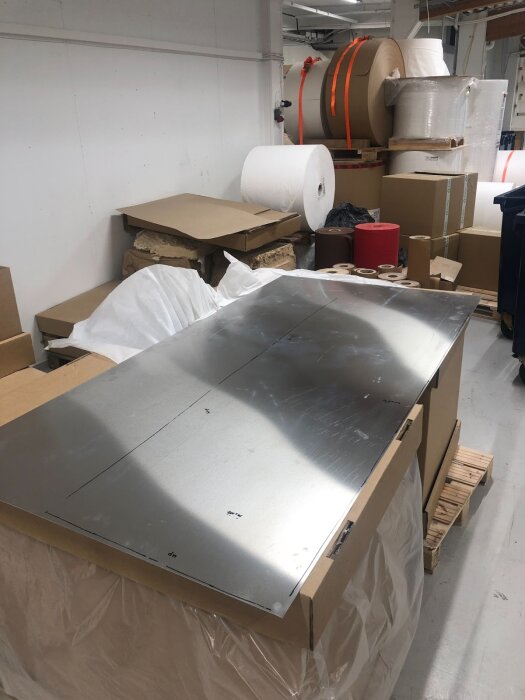 Aluminiumplåt på arbetsbord, för dörrtillverkning, omgiven av kartonger och stora pappersrullar i en verkstadsmiljö.