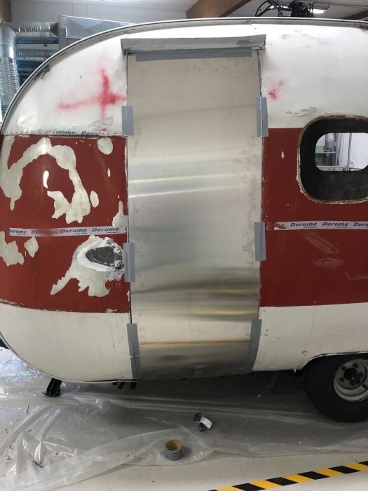 Aluminiumplåt tejpad över dörröppning på röd och vit husvagn under renovering i verkstad.