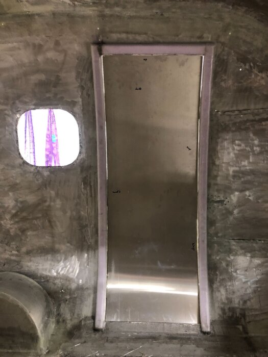 Nybyggd dörr med aluminiumpanel i en grå dörröppning under konstruktion, med fönster till vänster och lila gardiner synliga genom fönstret.