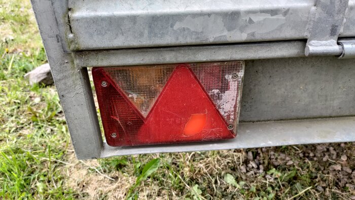 Trasigt bakljusglas på ett fordon, synligt rött och orange med en spricka på en grå bakgrund av metall. Gräs och stenar i bakgrunden.