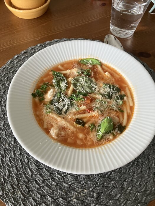 En tallrik minestronesoppa med bucatinipasta, toppad med spenat och riven parmesanost, serverad på ett bord med ett glas vatten.