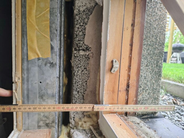 Närbild av källarfönster med öppen vägg, visar isolering, stålregel, och murad Leca, med tumstock som mäter avstånd till utsidan.