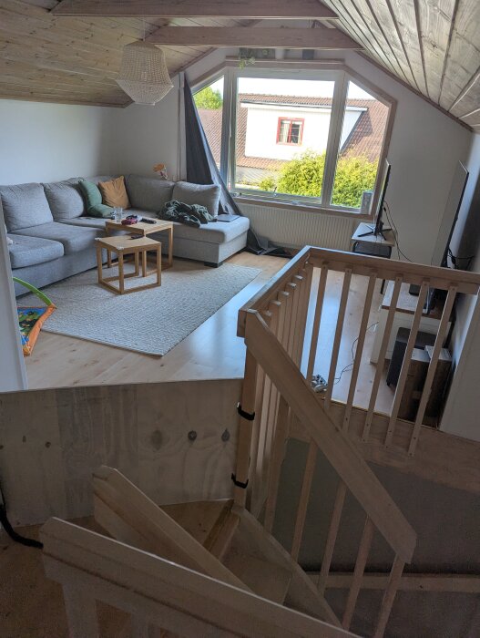 Trappa som leder upp till ett vardagsrum under snedtak med stor soffa, soffbord, stort fönster och TV, mörkläggningsgardin delvis öppen.