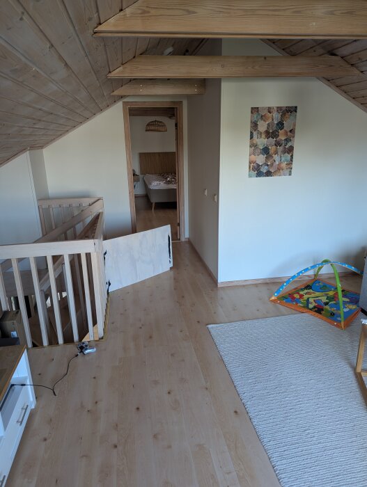Trappa och hall på övervåning i radhus med trägolv, sett mot sovrumsdörr. Inredning inkluderar babyhage och lekmatta, med snedtak och synliga takbjälkar.