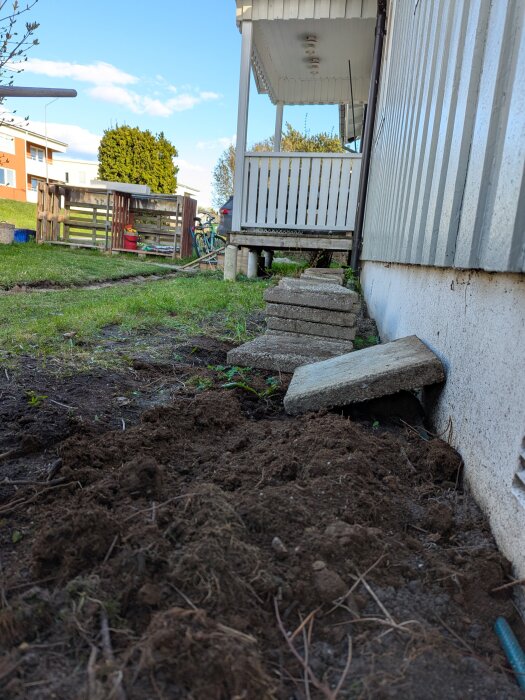 Uppstaplade stenplattor mot en husvägg, förberett med jord för att justera tomtens lutning, med en veranda i bakgrunden.