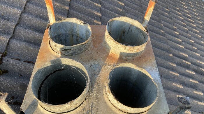 Fyra runda metallskorstenar på ett tak, omgivna av tegelpannor, med viss rost och spindelväv.