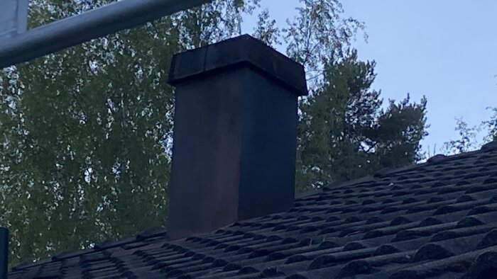 En skorsten med skyddshuv på ett tegeltak omgiven av träd.
