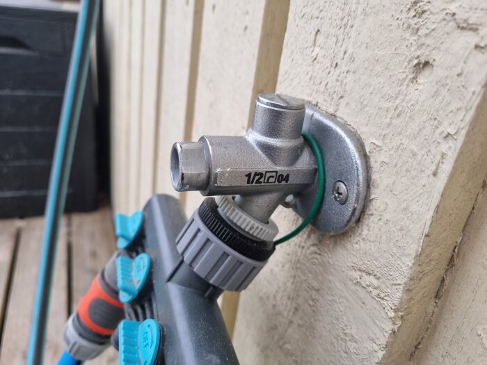 Frostsäker vattenutkastare utan synligt märke monterad på vit trävägg med slangkopplingar anslutna. Markering "1/2 04" synlig på ventilen.