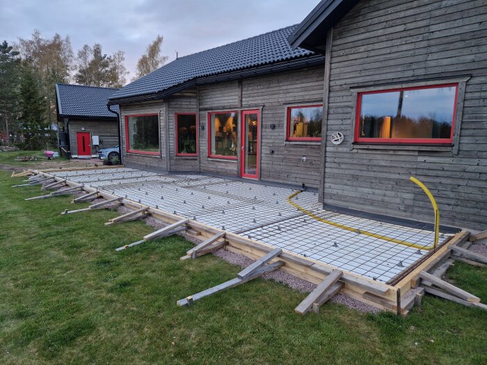 Förberedd grund för gjutning av altan och entréer vid ett trähus, med armeringsnät och trästöd, omgiven av gräsmatta.