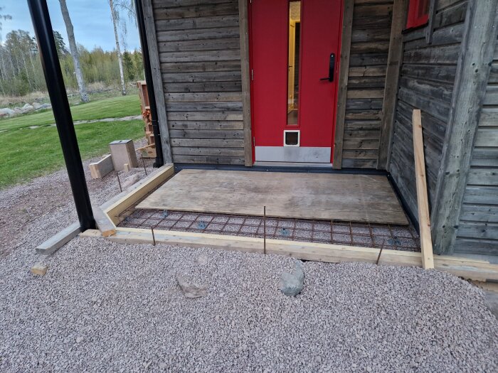 Form för gjutning av altan och entre framför en röd dörr på en träbyggnad, omgiven av grus och träplankor.
