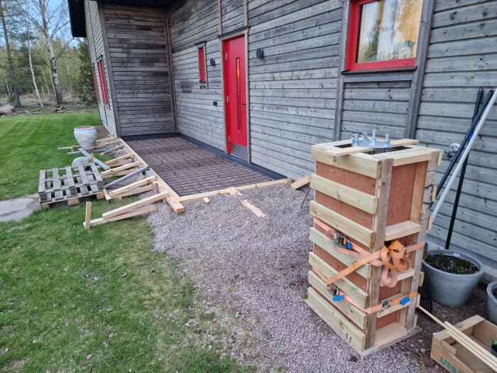 En träform för gjutning av altan och entréer vid ett hus med röd dörr, omgiven av grus och grönska. Flaggstångsfundament förberett.