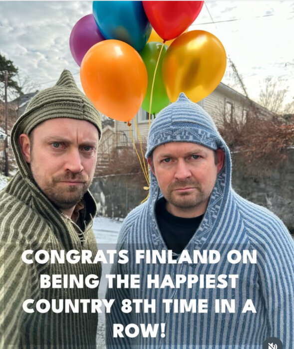 Två personer i stickade luvtröjor står framför ballonger i olika färger med text som gratulerar Finland till att vara det lyckligaste landet.
