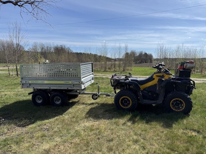 ATV kopplad till en Smålandsvagnen tippsläp på en gräsmatta, omgiven av träd och klar himmel i bakgrunden.