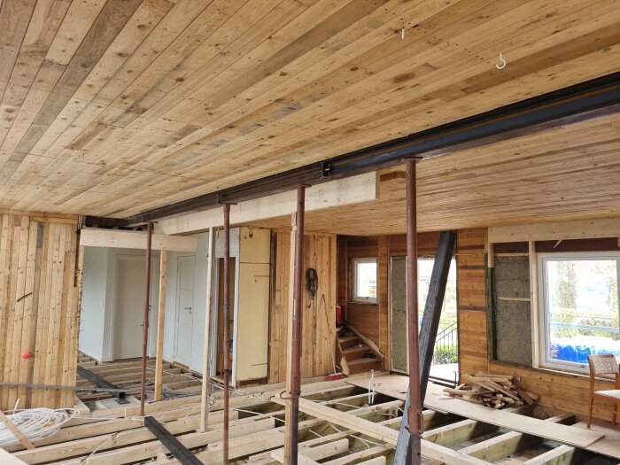 Byggarbetsplats med träväggar och tak, stålbalkar och pelare installerade, golv under konstruktion, fönster i bakgrunden.